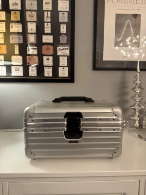 resväska Rimowa (Vanity case) aluminium - Säljer en ikonisk silverfärgad resväska från Rimowa i aluminium med robust design och svarta detaljer. Väskan har ett praktiskt handtag, tydliga nitar och ett smart inre fack med spegel. Perfekt för dig som vill resa med stil och ha koll på dina grejer.