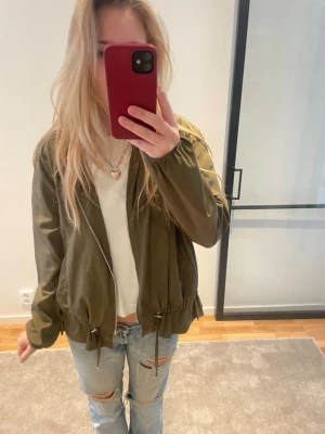 Olivgrön vindjacka med snörning - Säljer en olivgrön vindjacka med dragkedja framtill och snörning i midjan. Jackan har långärmade ärmar och en loose passform Materialet är lätt och syntetiskt, Från Zara och i storlek S. Superbra skick