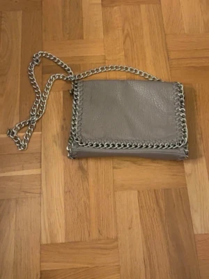 Beige axelväska från Tiamo - Säljer en äkta axelväska från Tiamo med grov silverfärgad kedja som axelrem och runt kanterna. Väskan är i skinnimitation och har magnetlås samt flera fack inuti. Väskan har dessvärre tappat lite av materialet, se bild 5.