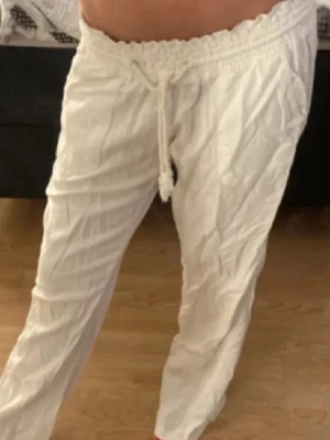 Vita linnebyxor med snörning - Säljer ett par luftiga vita linnebyxor med dragsko i midjan och raka ben. Byxorna har elastisk midja och fickor på sidorna. Perfekta för varma dagar och ger en avslappnad vibe. Dom har gått sönder lite vid ena snöret som man ser på bilden men inget man tänker på när man har på dom