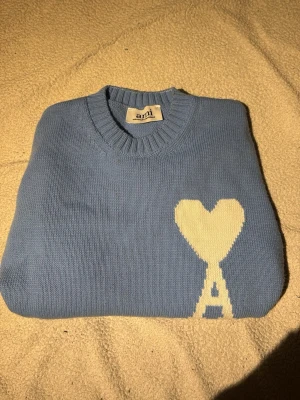 Ljusblå stickad tröja från Ami - Säljer en ljusblå stickad tröja från Ami med ikoniskt hjärta och bokstaven A i vitt på bröstet. Tröjan har rund hals, ribbade muddar och är långärmad. Perfekt för dig som gillar stilrena och trendiga plagg.