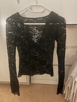 Svart spetsblus från Gina Tricot - Säljer en svart långärmad spetsblus från Gina Tricot i storlek XS. Blusen är helt genombruten med blommigt spetsmönster och har en djup v-ringning. Perfekt att styla med ett linne under eller som den är för en edgy look.