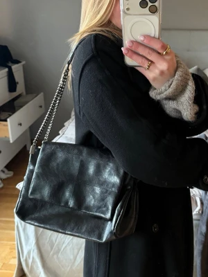 Svart väska med axelband från Zara - Säljer en svart axelväska från Zara i skinnimitation. Den har kedjeremmar i metall, flera fack, bland annat ett mindre med fack och tre större. Perfekt storlek för det viktigaste både för vardag och fest. Väldigt snygg detalj till en tråkig outfit. Hör gärna av dig vid frågor💗