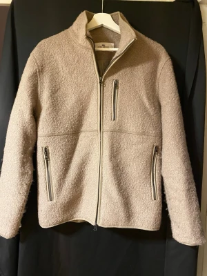 Beige fleccejacka från John Henric - Säljer en beige teddyjacka från John Henric i storlek M. Jackan har hög krage, tre fickor med dragkedja och en mjuk, fluffig yta. Perfekt för dig som vill ha en stilren och mysig jacka med snygga detaljer.