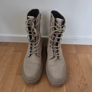 Beige mockaboots med snörning - Säljer ett par snygga beige boots i mockaliknande material med grov platt sula och rund tå. Skorna har snörning framtill och dragkedja på sidan för enkel på- och avtagning. Perfekta för dig som gillar chunky stil och vill ha något som sticker ut i höst och vinter. Använda någon enstaka gång.