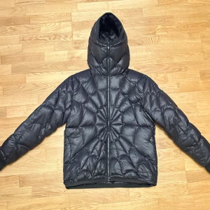 Moncler spider man  - Snygg svart dunjacka från Moncler med coolt quiltat mönster och stor huva. Jackan har dragkedja framtill, diskret Moncler-logga och ser riktigt varm ut. Materialet är glansigt och lätt, perfekt för kalla dagar. Insidan har en unik serietidningsetikett.