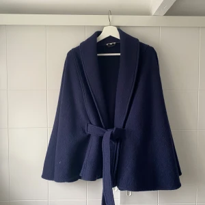 Poncho ullkappa med bälte - Säljer min fina mörkblå poncho kappa i ull med knytskärp i midjan. Kappan är från märket stenströms och har knappt används. Storleken är i L men tycker att den är mer som en s i storleken då den sitter perfekt på mig som bär xs i vanliga fall.   
