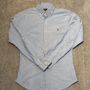 Ljusblå skjorta från Ralph Lauren - Säljer en stilren ljusblå skjorta från Ralph Lauren i slim fit. Skjortan har klassisk krage, knappar hela vägen och den ikoniska broderade polospelaren på bröstet. Perfekt för dig som gillar en clean och tidlös look.