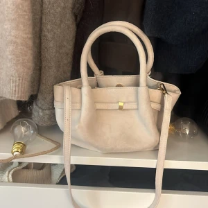 Ljusbeige handväska med axelrem - Snygg ljusbeige handväska i mocka-look med både handtag och justerbar axelrem. Väskan har guldfärgade metalldetaljer och en stilren, minimalistisk design. Perfekt storlek för det viktigaste och passar till många outfits.