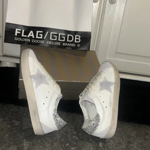 Golden Goose vita sneakers med glitter - Golden Goose sneakers i vitt skinn med grå mockadetaljer och silverglittrig häl. Klassisk stjärna på sidan, snörning framtill och platt sula. Snygg kontrast mellan det matta och glittriga, perfekt för dig som gillar att sticka ut.