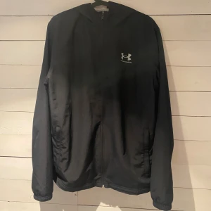 Svart vindjacka från Under Armour M - Svart vindjacka från Under Armour med huva och dragkedja framtill. Jackan har en loose passform och är tillverkad i lätt polyester med meshfoder för extra komfort. Perfekt för dig som vill ha en sportig och stilren look.