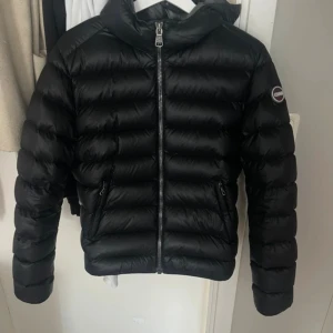 Svart dunjacka från Colmar - Snygg svart dunjacka från Colmar med glansig finish och horisontella sömmar. Jackan har huva, två dragkedjeförsedda fickor och en tydlig logga på ärmen. Perfekt för kalla dagar och riktigt skön att bära. Stängs med dragkedja framtill. Inga defekter! Skriv vid frågor💕