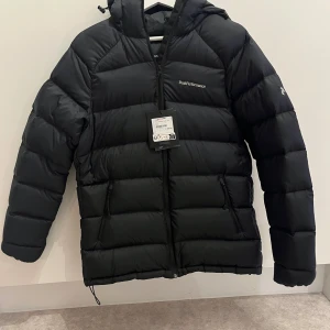 Svart dunjacka Peak Performance M - Peak Performance svart dunjacka ny alldrig använd med huva och dragkedja. Jackan är quiltad och har två sidofickor med dragkedja. Perfekt för kalla dagar och har en clean, sportig look. Broderad logga på bröstet och ärmen. Modellen heter W Down Hood Jacket. Säljes pga fel köp