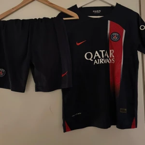 PSG matchställ Nike blå/röd - Paris Saint-Germain matchställ med kortärmad tröja och shorts. Tröjan är marinblå med röd och vit rand, PSG-logga på bröstet och Nike-logga i rött. Materialet är lätt och andas, perfekt för fotbollsträning eller match.