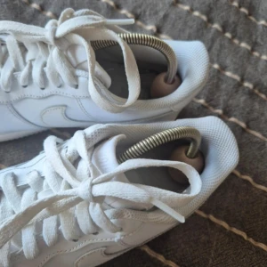 Nike Air Force 1 vita sneakers st 39 - Klassiska Nike Air Force 1 sneakers i helvitt skinn med rund tå och snörning. Snygg och tidlös modell med perforerad tåbox och ikonisk swoosh-logga på sidan. Perfekta för dig som vill ha en clean och stilren look till vardags.