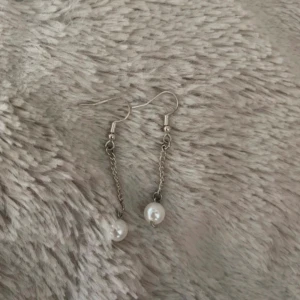 Silvriga örhängen med pärldetalj - Säljer ett par stilrena örhängen i silverfärg med tunn kedja och vit pärla längst ner. Perfekta för dig som gillar minimalistiska smycken med en elegant touch. Lätta att bära och passar till många olika looks.
