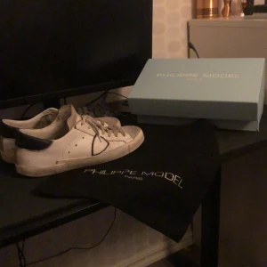 Philippe Model vita sneakers - Säljer ett par stilrena vita sneakers från Philippe Model Paris med mörkblå häl och klassisk snörning. Skorna har en rund tå och platt sula, tillverkade i skinn. Levereras med originalkartong och dustbag. Perfekt för dig som gillar exklusiva sneakers med fransk vibe.