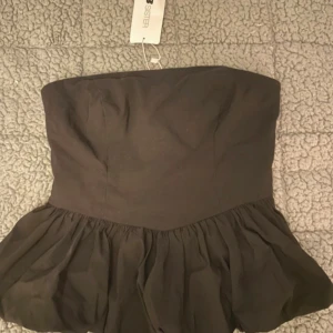 Svart axelbandslös kort topp  - Supersnygg svart axelbandslös kort topp från New yorker med volangkjol och markerad midja. Klänningen har dragkedja i sidan och smock baktill för extra komfort. Perfekt för dig som vill ha en stilren och trendig look.