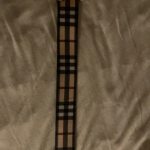 Burberry rutig skärp med guldspänne - Snyggt Burberry-bälte i klassiskt rutigt mönster med beige, svart, vitt och röda detaljer. Bältet har ett exklusivt guldigt TB-spänne och är gjort i äkta skinn. Perfekt accessoar för att lyfta din outfit med en lyxig touch.