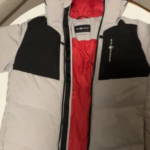 Sail Racing dunjacka i grått och svart - Säljer en fet dunjacka från Sail Racing i grått med svarta detaljer och röd insida. Jackan har huva, två bröstfickor med dragkedja och logga på ena sidan. Perfekt för kalla dagar och har en clean, sportig vibe.