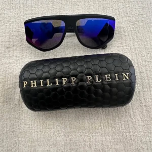 Philipp Plein svarta solglasögon - Snygga svarta solglasögon från Philipp Plein med spegelblå-lila glas och bred båge. Ikonisk PP-logga på sidan och coolt, sportigt uttryck. Kommer med ett svart fodral i hexagonmönstrat material med guldtext. Perfekta för dig som vill sticka ut.