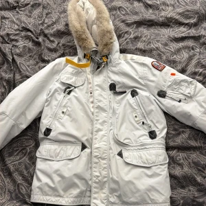 Vit Parajumpers vinterjacka med päls - Säljer en vit Parajumpers vinterjacka, modell Anchorage, med päls på huvan och ikonisk gul rem. Jackan har flera fickor, patch på ärmen och dragkedja framtill. Slim fit och riktigt varm, perfekt för kalla dagar.
