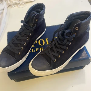 Mörkblå high top sneakers Polo Ralph Lauren - Snygga mörkblå high top sneakers från Polo Ralph Lauren med vit sula, guldfärgade öljetter och detaljer i mocka och skinn. Klassisk design med snörning och Polo-logga på hälen. Perfekta för dig som vill ha en stilren och sportig look.