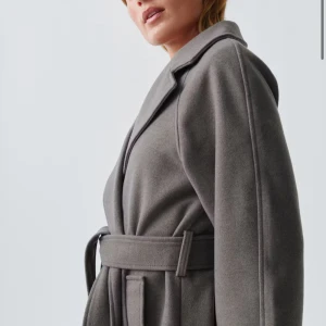 Grå kappa med bälte  - Säljer en stilren grå kappa med breda slag, omlottmodell och knytbälte i midjan. Kappan har lång ärm och en rak, loose passform som ger en chill vibe. Materialet är mjukt och känns lyxigt, perfekt för kalla dagar. 