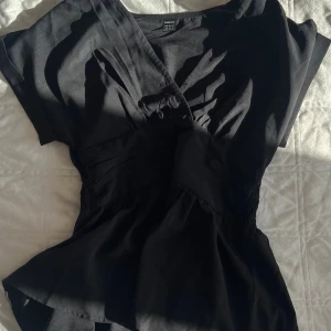 Svart omlottopp från SHEIN XS - Svart omlottopp från SHEIN i storlek XS. Toppen har v-ringning, korta ärmar och markerad midja med rynkade detaljer. Snygg och enkel modell som ger en feminin siluett. Perfekt att styla med jeans eller kjol.