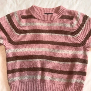 Randig rosa stickad tröja - Superfin stickad tröja i rosa med bruna och vita ränder. Tröjan har ribbad halsringning och långa ärmar. Mjuk och skön känsla med en klassisk passform. 