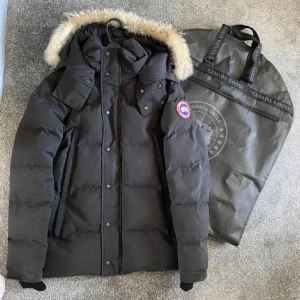 Svart dunjacka från Canada Goose - Canada Goose Dunjacka – Svart med Äkta Päls  Säljer en svart Canada Goose-dunjacka med äkta päls på huvan. Jackan har den klassiska pufferdesignen, broderad logga på ärmen och stora fickor framtill. Tillverkad i högkvalitativa material som ger utmärkt värme och komfort under kalla vinterdagar.  Ett exklusivt och tidlöst plagg som kombinerar funktionalitet, lyx och modern streetstil.