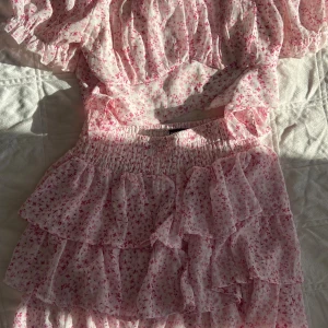 Rosa blommig volangkjol och topp - Säljer en söt kort kjol och en topp med rosa blommigt mönster och flera lager volanger. Kjolen har smock i midjan för skön passform och är gjord i ett lätt, luftigt material som passar perfekt till varma dagar. Supersöt och feminin vibe! Jag säljer både toppen och kjolen tillsammans för endast 225 kr !!!