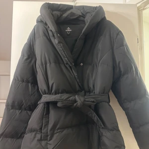 Svart pufferjacka från Threadbare - Snygg svart pufferjacka från Threadbare med stor huva och bälte i midjan. Jackan har quiltad design, knappar framtill och en oversized look som ger en cool vibe. Perfekt för kalla dagar när du vill vara både varm och stilren.