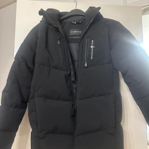Svart pufferjacka från Sail Racing - Svart pufferjacka från Sail Racing med huva och broderad logga på ryggen. Jackan har två bröstfickor med dragkedja och en snygg, stilren design. Perfekt för kalla dagar och riktigt skön att bära. Materialet är slitstarkt och håller värmen bra.
