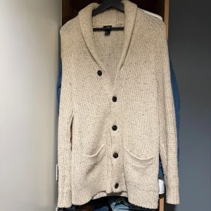 Stickad cardigan - Beige stickad cardigan från H&M med knappar och två fickor framtill. Koftan har bred krage och bruna mockadetaljer på armbågarna. Perfekt för lager-på-lager under höst och vinter. Sitter stort i storlek tycker jag