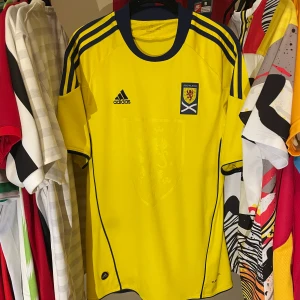 Skottland gul fotbollströja Adidas 2011/12 äkta Strl M - Gul fotbollströja från Skottlands landslag säsöngen 2011-2012, Adidas med blå detaljer och ränder på axlarna. Tröjan är i bra skick utöver en liten missfärgning på framsidan, Tröjan är i storlek medium,  trots att storleksetiketten är sliten bedömmer jag att den är i just M.