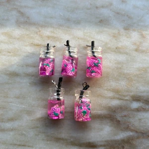 Berlocker med rosa bär i glasflaska - Unika berlocker formade som små glasflaskor fyllda med rosa bär och svarta sugrör. Färgglada detaljer i rosa, grönt och vitt ger en lekfull och kreativ look. Perfekta för dig som vill sticka ut med ett annorlunda smycke.