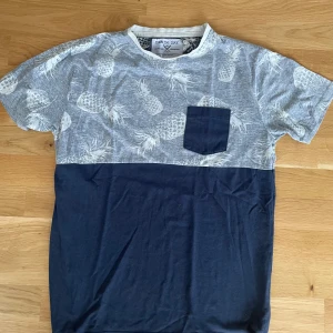 Gråblå t-shirt med ananas från Denim Day - Snygg t-shirt från Denim Day i grått och mörkblått. Överdelen har ett vitt ananasmönster och en mörkblå bröstficka. T-shirten har rund halsringning med vit kant och är kortärmad. Perfekt för dig som gillar coola prints och kontraster.