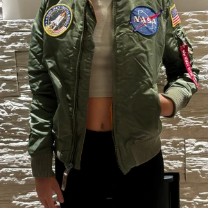 Grön bomberjacka NASA Alpha Industries - Säljer en grön bomberjacka från Alpha Industries med NASA-patchar och amerikansk flagga på ärmen. Jackan har orange foder, dragkedja framtill och ribbade muddar. Klassisk pilotstil med coola detaljer och ficka på ärmen.