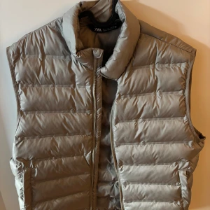 Beige dunväst från Zara - Snygg beige dunväst från Zara med quiltad design och hög krage. Västen har dragkedja framtill och praktiska fickor på sidorna. Perfekt för lager-på-lager och ger en clean look till din outfit.