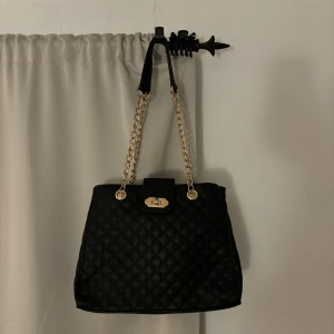 Svart quiltad axelväska med kedja - Stilren svart axelväska med quiltat mönster och snygga guldfärgade kedjehandtag. Väskan har en rektangulär form, lock med vridlås i guld och är tillverkad i skinnimitation. Perfekt för dig som gillar klassisk design med en modern twist. Lite sliten men inget som syns 