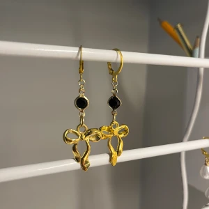 Guldiga örhängen med rosett och svart sten - Unika örhängen i guldton med hängande rosettformad detalj och en svart sten ovanför. Perfekt accessoar för dig som vill sticka ut med något sött och elegant. Säljs som ett par. Handgjort