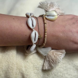 2 Armband med snäckor och tofsar - Säljer ett set med 2 armband i avslappna boho-stil, dekorerade med vita snäckor, små träpärlor och stora tofsar i ljusbeige. Armbanden har även en liten i guldton och är perfekta för en somrig look. Reglerbar storlek. 