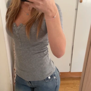 Grå ribbad topp med knappar - Säljer en grå ribbad topp med v-ringning och flera knappar framtill. Toppen har korta ärmar och sitter tight mot kroppen,väldigt vacker och ny 
