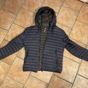 dunjacka från Colmar - Snygg mörkblå dunjacka från Colmar med huva och horisontella sömmar. Jackan har dragkedja framtill, två sidofickor med dragkedja och en lätt, quiltad känsla. Perfekt för kyliga dagar när du vill hålla stilen enkel och clean.