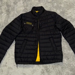 Svart Barbour jacka - Säljer en svart pufferjacka från Barbour International med gul insida och coolt bröstfack med logga. Jackan har dragkedja, ståkrage och flera fickor. Perfekt för dig som vill ha en lätt och snygg jacka med streetkänsla. Perfekt inför hösten, använd 2 gånger, så gott som nytt