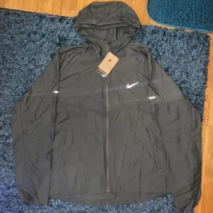 Grå Nike Miller windbreaker  - Svart vindjacka från Nike med huva och reflekterande detaljer. Jackan har dragkedja framtill, två fickor med dragkedja och en stilren Nike-logga på bröstet. Perfekt för blåsiga dagar och sportiga outfits.