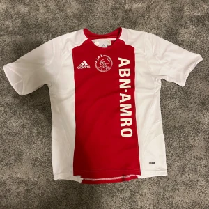 Ajax fotbollströja 2007/08 - Ajax matchtröja från Adidas i storlek 164. Klassisk design med röd mittpanel och vita sidor, samt tryckta loggor och sponsor ABN AMRO. Tillverkad i lätt och ventilerande ClimaCool-material, perfekt för träning eller match. Rund hals och korta ärmar.
