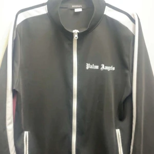 Svart track jacket från Palm Angels - Svart track jacket från Palm Angels med vita ränder längs ärmarna och vit dragkedja. Jackan har hög krage, fickor med dragkedja och logga på bröstet. Perfekt för en sportig och trendig look.