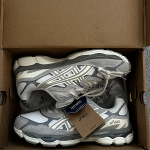 Asics GEL- NYC Cream/Oyster Grey - Säljer ett par fräscha Asics sneakers i grått och vitt med snygga detaljer. Skorna har meshpaneler för bra ventilation, syntetiska och mockaliknande partier samt en platt sula med dämpning. Klassisk Asics-logga på sidan och snörning framtill. Perfekt för dig som gillar stilrena och sportiga sneakers.